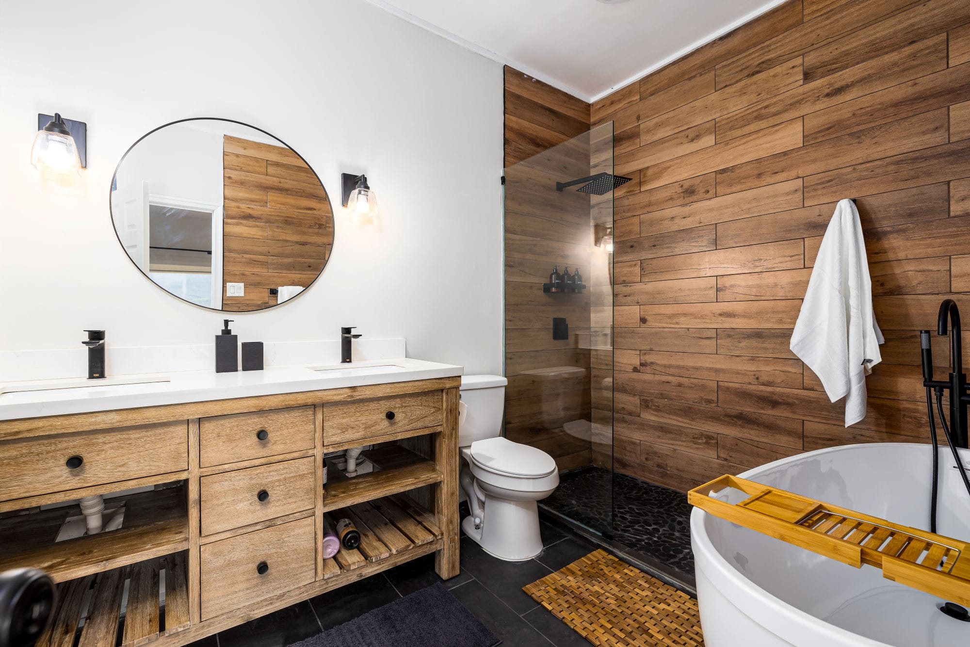 Spa bathroom with a rustic wood double vanity, round mirror, wood-tile walk-in shower, and a freestanding tub