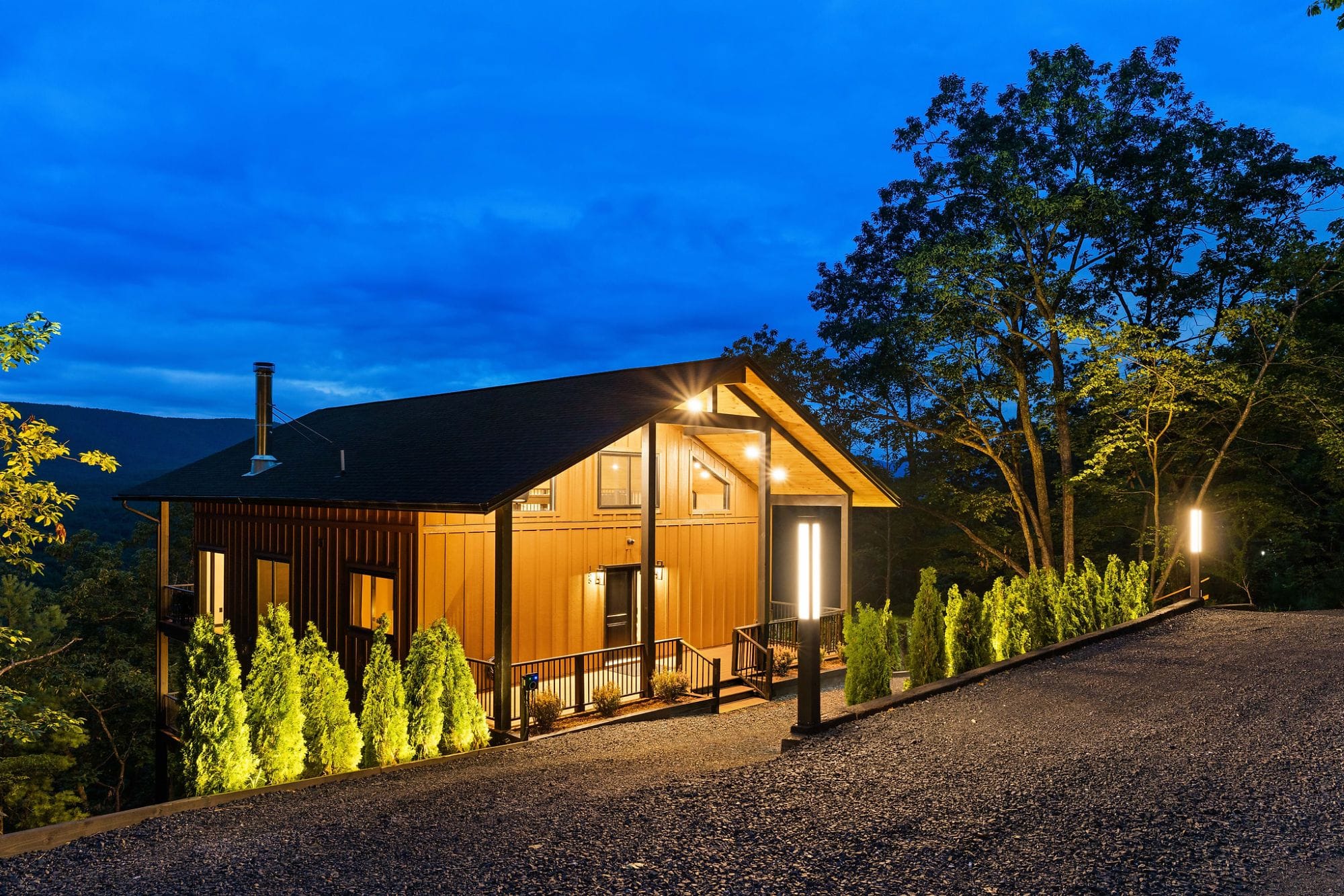 The Cabin at blue hour, warm interior glow with landscape-lit arborvitae along the gravel drive